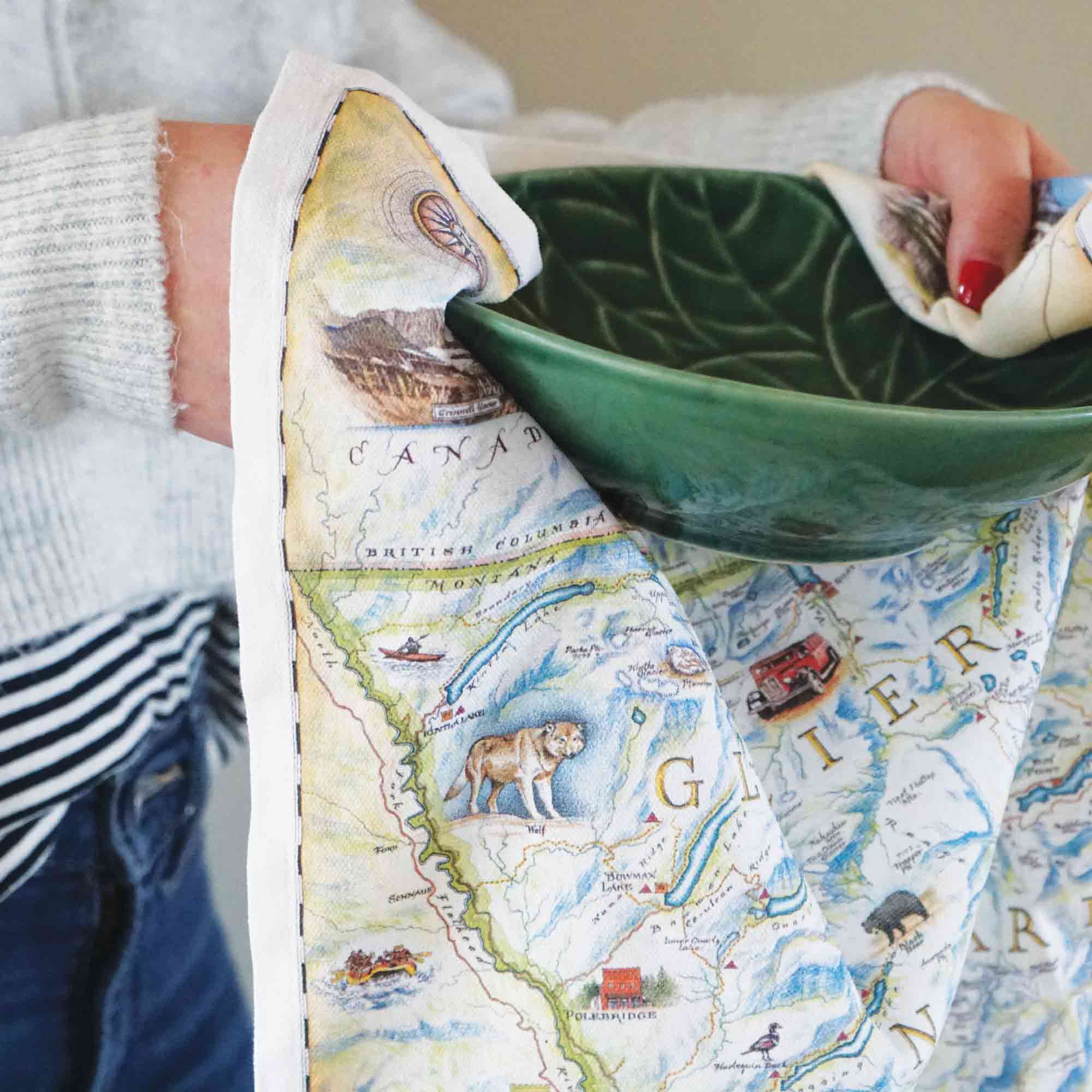 A women drying a green bowl with a Glacier Kitchen Dishwashing Towel. The map features places in the park such as Logan Pass, Lake McDonald Lodge, Many Glacier Lodge, Two Medicine, Glacier Park Lodge, and the Going-to-the-sun road. Flora and fauna include grizzly bear, elk, moose, mountain goat, and mountain lion.