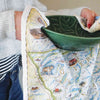 A women drying a green bowl with a Glacier Kitchen Dishwashing Towel. The map features places in the park such as Logan Pass, Lake McDonald Lodge, Many Glacier Lodge, Two Medicine, Glacier Park Lodge, and the Going-to-the-sun road. Flora and fauna include grizzly bear, elk, moose, mountain goat, and mountain lion.