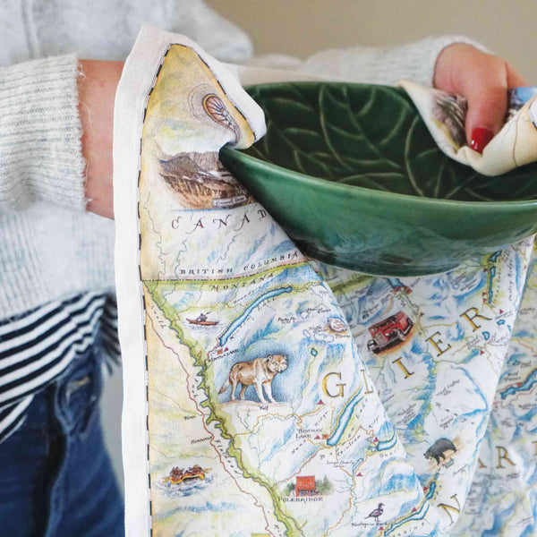 A women drying a green bowl with a Glacier Kitchen Dishwashing Towel. The map features places in the park such as Logan Pass, Lake McDonald Lodge, Many Glacier Lodge, Two Medicine, Glacier Park Lodge, and the Going-to-the-sun road. Flora and fauna include grizzly bear, elk, moose, mountain goat, and mountain lion.
