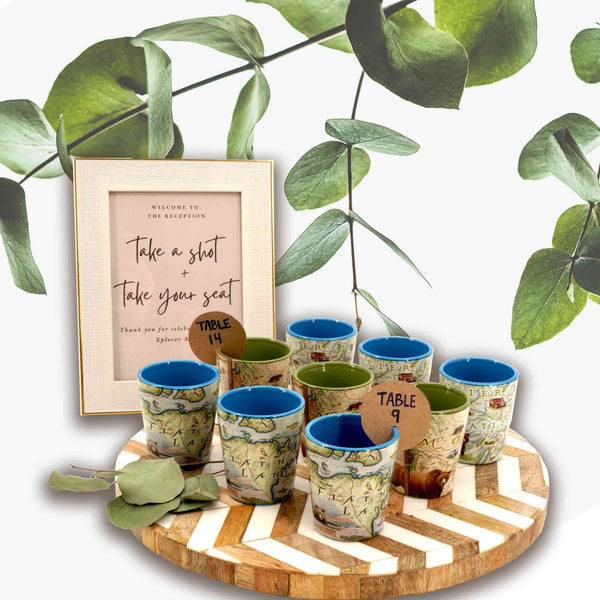 Nine shot glasses sitting on a white and wood round tray. There is a sign behind it that says, "Take a Shot and Take your seat." The shot glasses have table numbers on a brown tag.