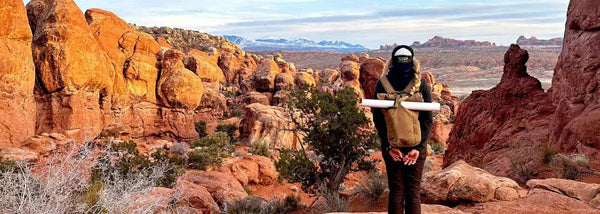 A women standing over a canyon cliff looking at into Arches National Park. She has a rolled up map in her backpack flap.