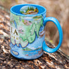 Maine's Acadia National Park Map Ceramic Mug in the color blue sitting on a log. The coffee cup features Cadillac Mountains, Otter Cliffs, Beaver, and Jordon Pond.