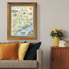 Xplorer Maps state map of Maine hangs in a living room above a gray couch with orange pillows.