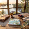 A square ceramic tile featuring a colorful vintage map of '1900 Islands' is displayed on a rustic wooden table. The scene is styled with a leather-bound notebook, a brass compass, a bowl of cherries and blueberries, and a folded linen cloth, all set against a large window with a scenic view of a lake and forest at sunset.
