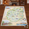 A completed jigsaw puzzle of The Adirondacks map spread on a wooden table, with a few loose pieces nearby and a blurred cozy indoor room in the background.