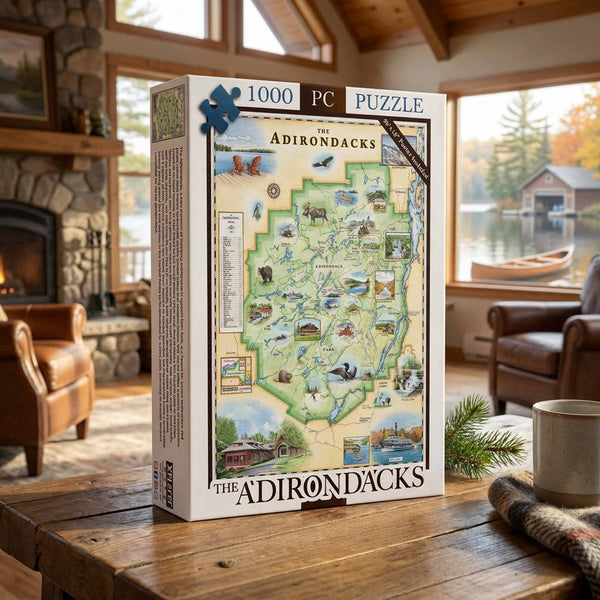 A 1000-piece jigsaw puzzle box titled 'The Adirondacks' stands on a rustic wooden table in a cozy living room setting with a stone fireplace, leather armchair, and window view of a lake and boat.