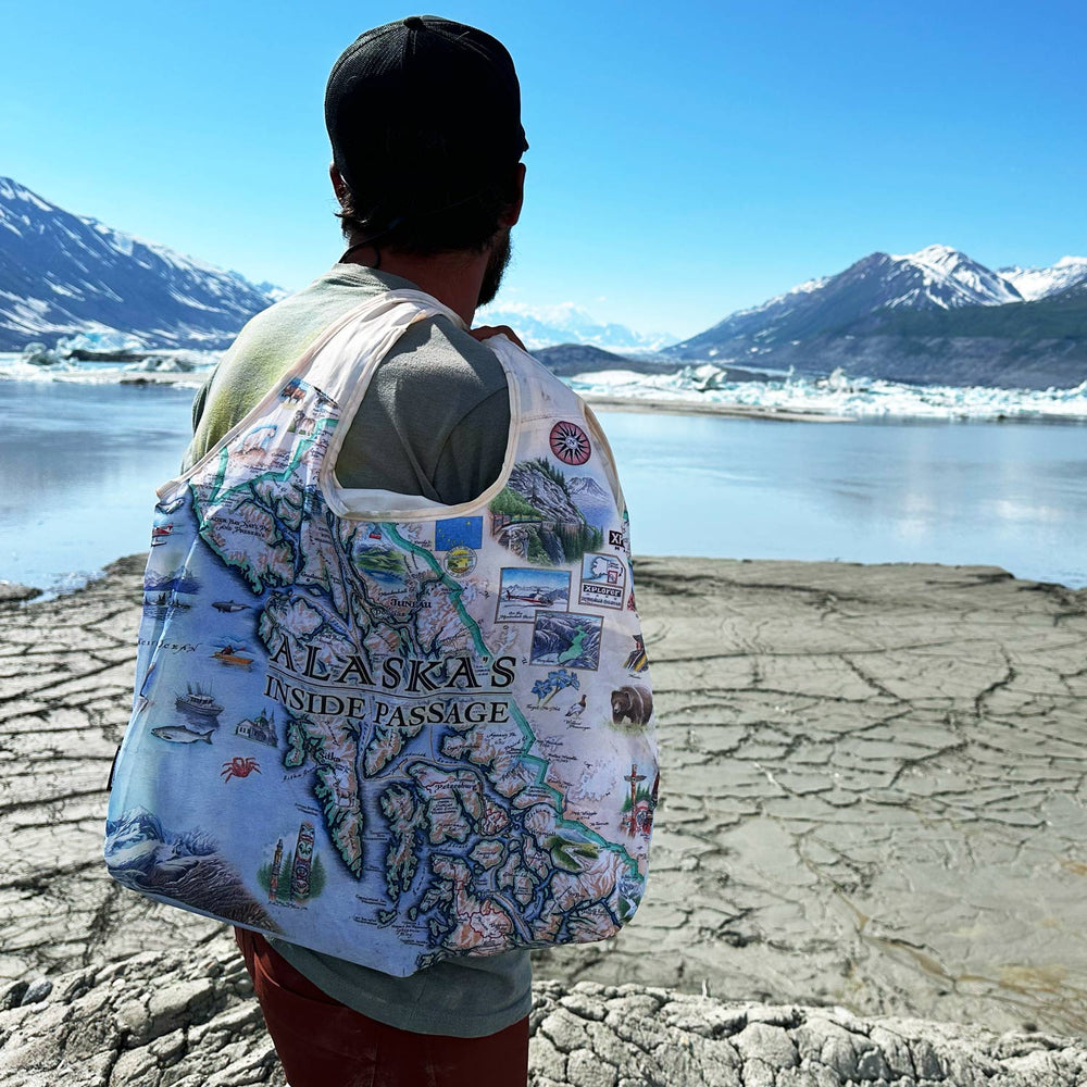 Hiker wearing a hooded jacket and backpack, carrying an Alaska's Inside Passage map pouch tote, with snow-capped mountains in the background.