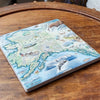 4" x 4" Alaska State map ceramic coaster in earth tone colors of blues and greens, sitting on a coffee table. The map includes Denali National Park and cities such as Anchorage, Fairbanks, and Juneau. Illustrations of wildlife matching the region: Polar b