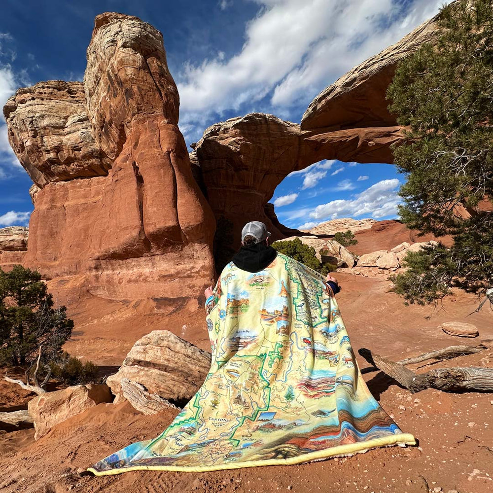 A Man with Utah's Arches & Canyonlands National Park fleece blanket wrapped around him. backdrop: Red Rock formation inside the national park. 