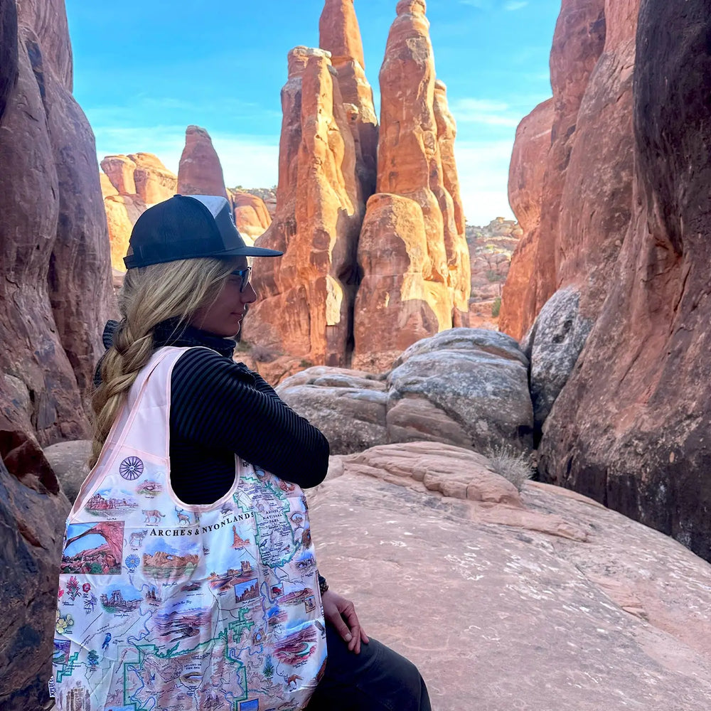 Woman wearing a hat carrying an Arches National Park map pouch tote bag, standing in front of scenic desert rock formations.