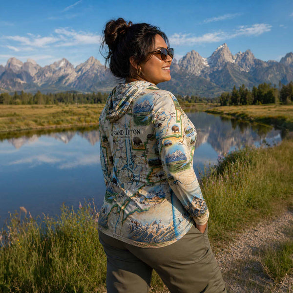 Woman in a scenic landscape with mountains and a lake, wearing a map-patterned sunshirt.