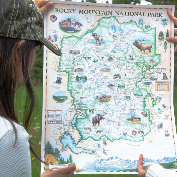 A person holding up a detailed illustrated map of Rocky Mountain National Park outdoors. The map features a green border, topographical line work, and numerous small illustrations of wildlife, lakes, forests, and landmarks.