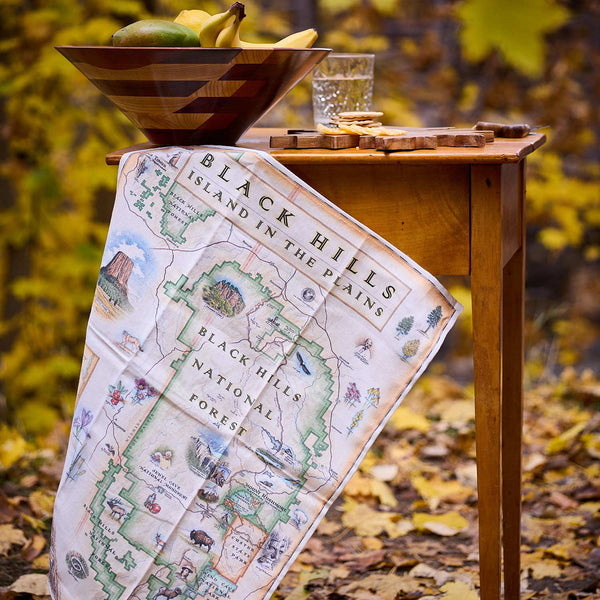 Map of Black Hills National Forest draped over a wooden table with a bowl of fruit and glass in an outdoor setting.