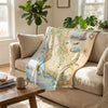 Beige sofa with a decorative blanket featuring a map design, in a room with plants and a coffee table.