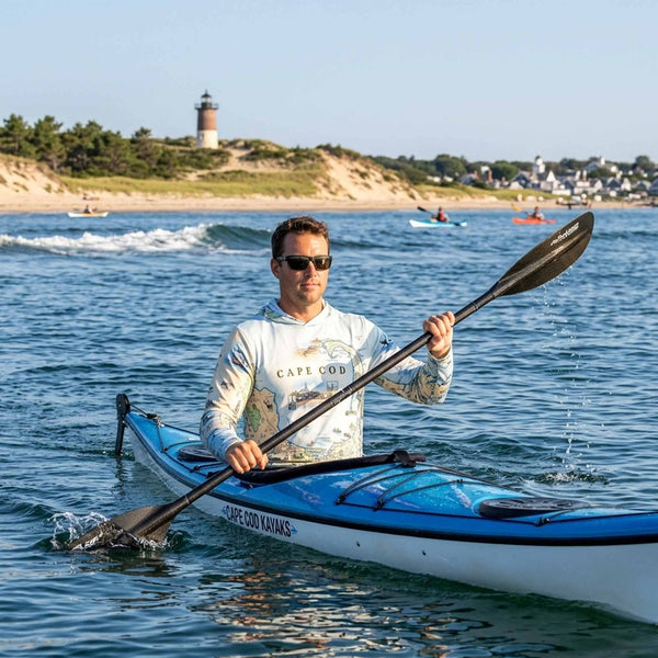 Man paddling a kayak on the water with a lighthouse in the background and wearing a Cape Cod sun shirt hoodie