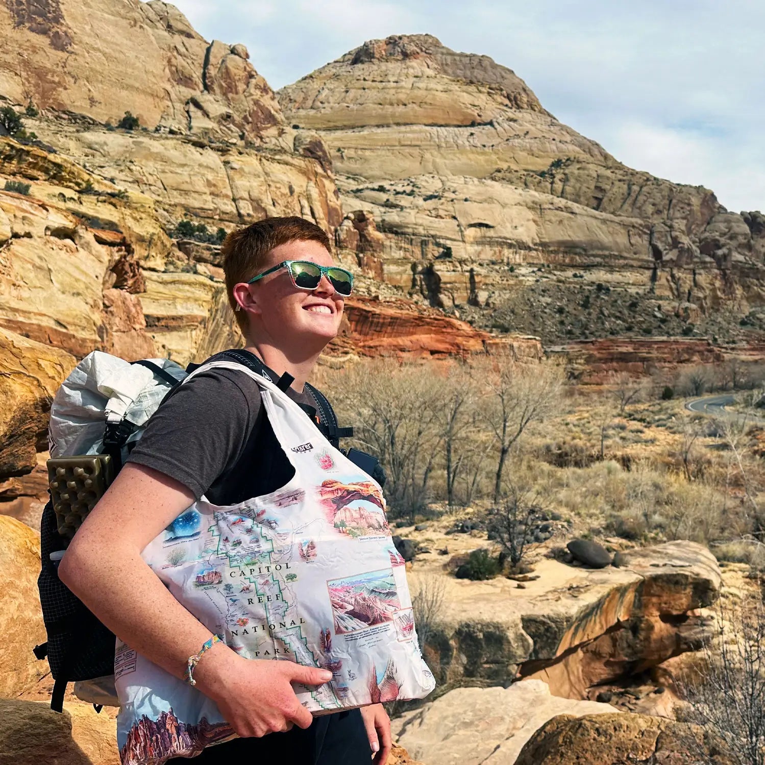 Person hiking in Capitol Reef National Park, wearing sunglasses and carrying a Capitol Reef National Park map pouch tote bag, with scenic desert rock formations in the background.