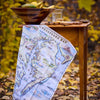 Map-themed towel draped over a wooden table with a bowl of fruit and glass in an autumn setting.