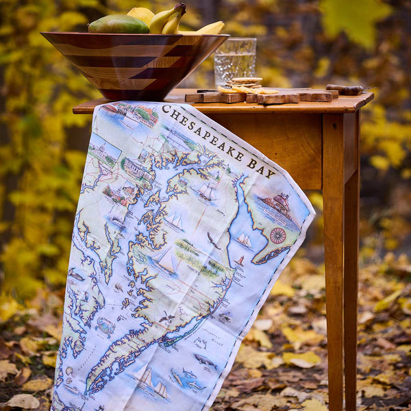 Map-themed towel draped over a wooden table with a bowl of fruit and glass in an autumn setting.