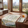 Dish towel with a map of Rocky Mountain National Park on a tablecloth, next to a glass of iced tea and a wooden spoon.