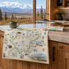 A vintage-style illustrated map print of Denali hangs over a wooden kitchen counter, with a matching rolled print and a mug nearby. A large window behind shows a snowy mountain landscape.