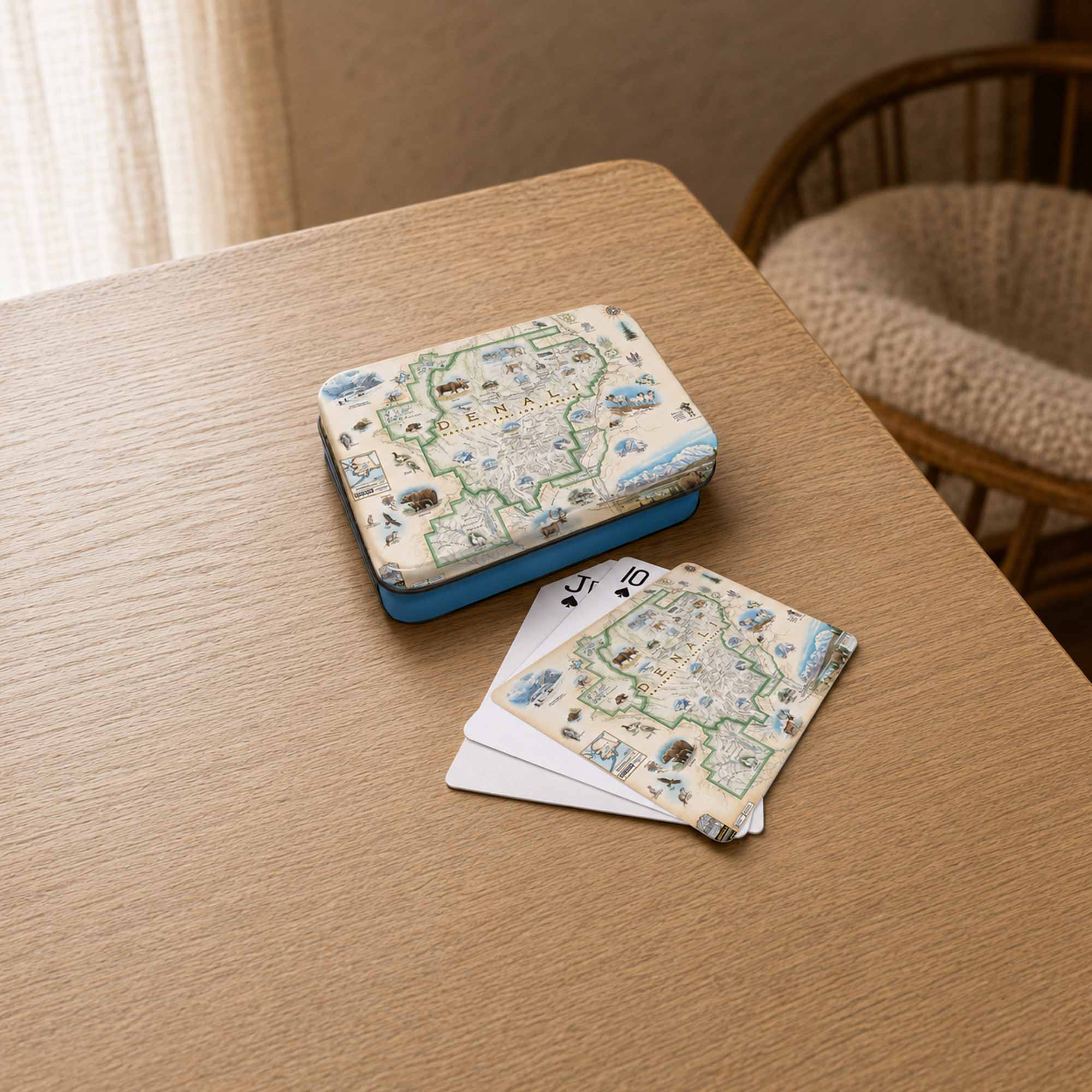 A deck of illustrated playing cards and its matching box resting on a light wood table, with a woven chair visible in the softly lit room background.