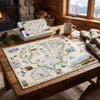 A completed jigsaw puzzle of a vintage map of Montana is displayed on a rustic wooden table in a cozy cabin setting, alongside its box, a mug, a knit sweater, and glasses.