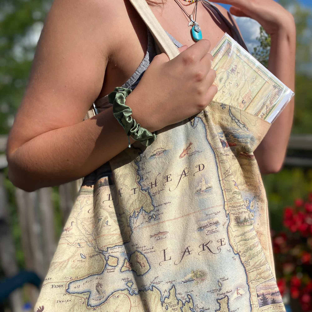 A woman carrying a Flathead Lake Montana Map Canvas Tote Bag featuring Golfing, Rafting, birds, eagles, Osprey, Bears, deer, mountain lion, fish, Cherry Blossoms, and flowers like Glacier Lilies and Lady Slippers.