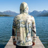 Person wearing a sun shirt hoodie with a Flathead Lake map design standing on a dock by a lake with mountains in the background