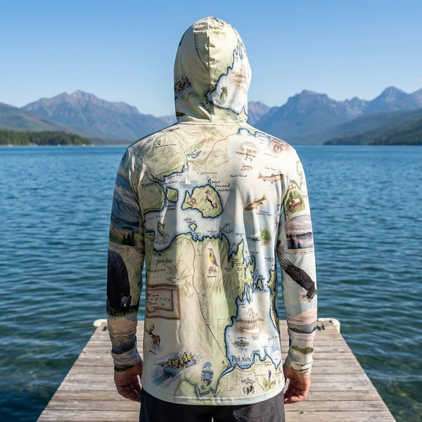 Person wearing a sun shirt hoodie with a Flathead Lake map design standing on a dock by a lake with mountains in the background