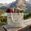 Map-patterned tote bag with camping gear against a mountainous landscape