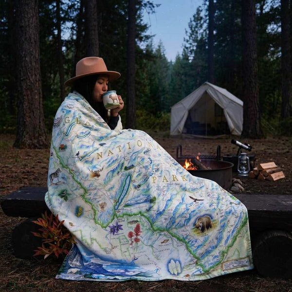 Person wrapped in a National Park map blanket by a campfire in the woods.