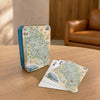 A deck of illustrated playing cards with a vintage map design stands on a wooden table next to a blue metal tin, with a few cards fanned out. A brown leather armchair and wood-paneled room are softly blurred in the background.