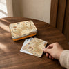 A hand holds a square playing card with an illustrated map design over a wooden table, next to a matching card tin and another card.