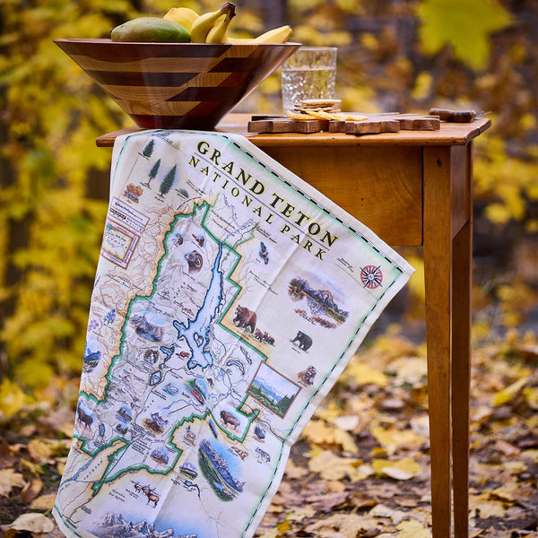 Map of Grand Teton National Park draped over a wooden table with a bowl of fruit and glass in an autumn setting.