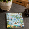 A square ceramic coaster featuring a colorful illustrated map of Hawaii, placed on a dark gray countertop next to a potted plant and a glass bottle.