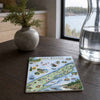 A square ceramic coaster featuring a colorful illustrated map of Isle Royale National Park, styled on a dark wooden table next to a small potted plant and a glass carafe.