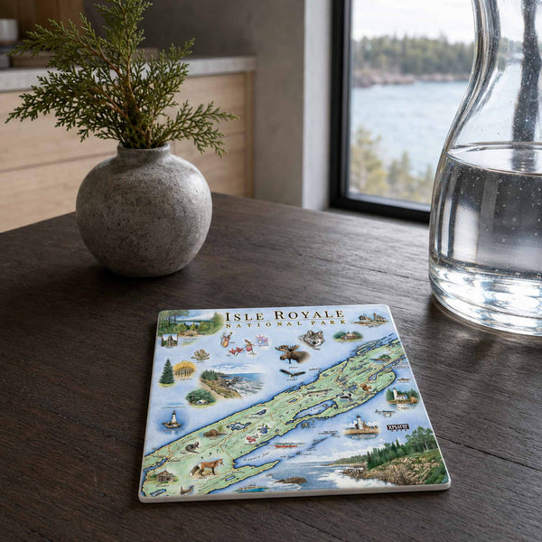 A square ceramic coaster featuring a colorful illustrated map of Isle Royale National Park, styled on a dark wooden table next to a small potted plant and a glass carafe.