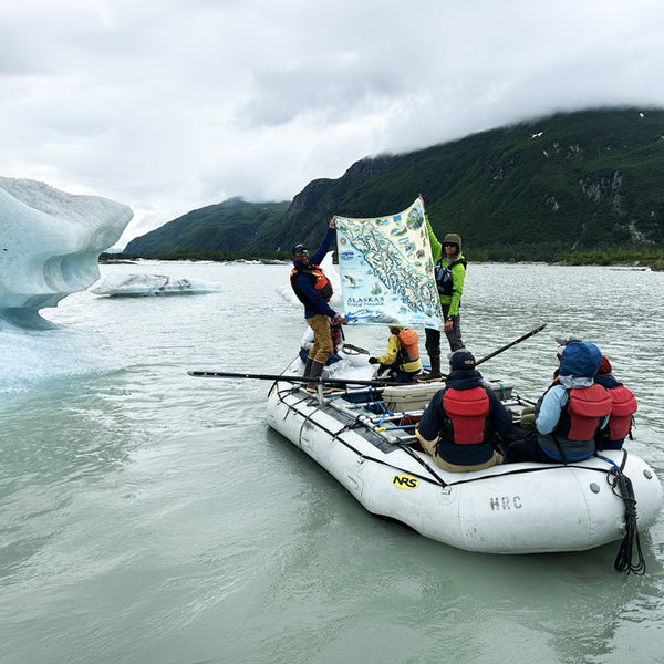 Group of people on a raft in a glacial lake with mountains in the background holding up an Inside Passage blanket
