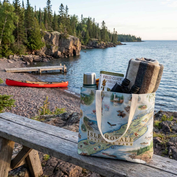 Tote bag with nature-themed design on a wooden bench by a lake with a red canoe and rocky shoreline.