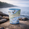 A light blue ceramic tumbler printed with a scenic Isle Royale National Park map and illustrations, set on a large rock with the ocean and shoreline blurred in the background.