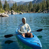 Person in a blue kayak on a calm Tahoe lake with mountains and trees in the background wearing a Lake Tahoe sun shirt hoodie