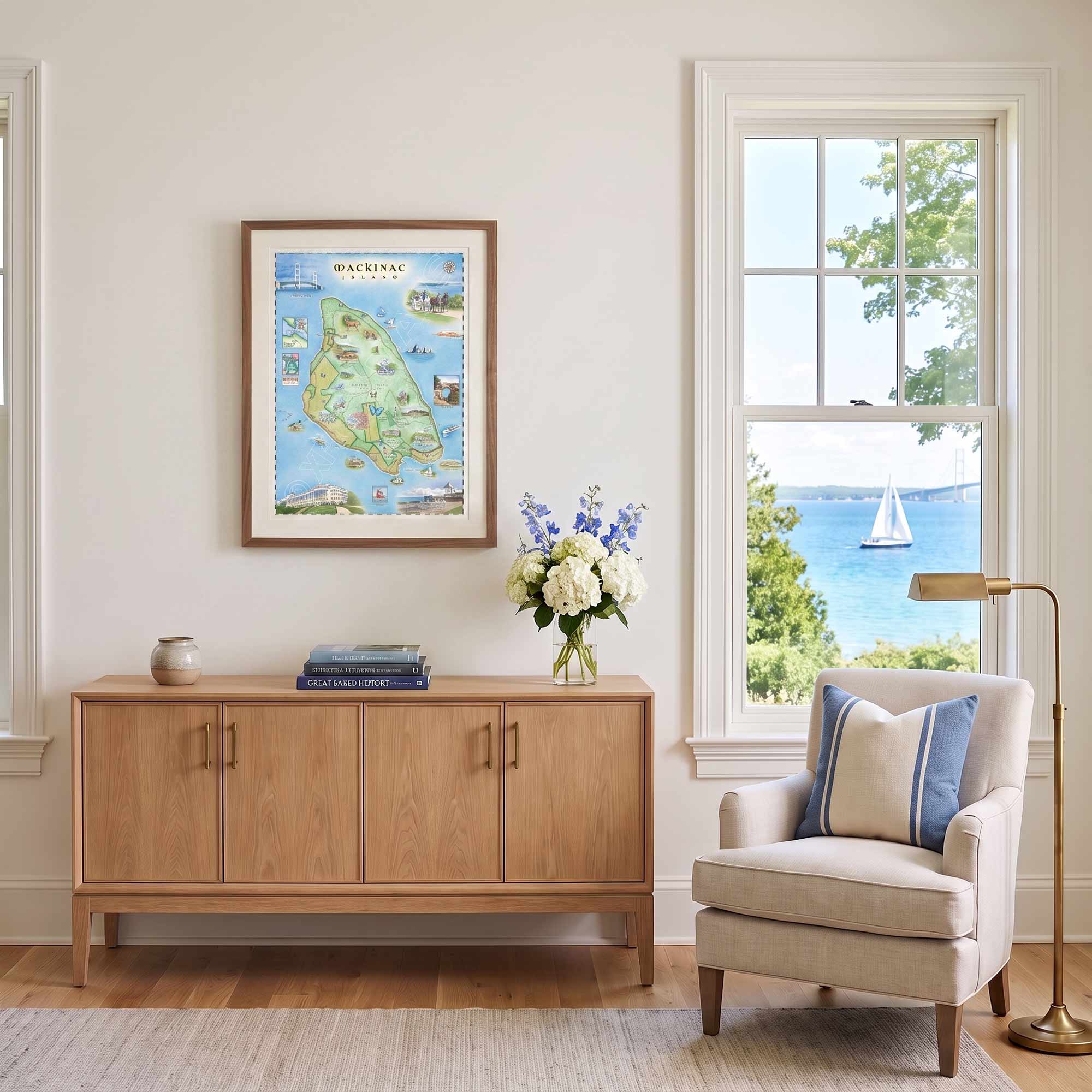 A framed art print of a colorful map of Martha's Vine Island, Virginia, hangs on a cream wall in a beautifully styled living room above a light wood credenza. The room features a beige armchair with blue and white striped pillows, a brass floor lamp, and a window looking out onto a scenic blue ocean with a sailboat.