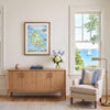 A framed art print of a colorful map of Martha's Vine Island, Virginia, hangs on a cream wall in a beautifully styled living room above a light wood credenza. The room features a beige armchair with blue and white striped pillows, a brass floor lamp, and a window looking out onto a scenic blue ocean with a sailboat.