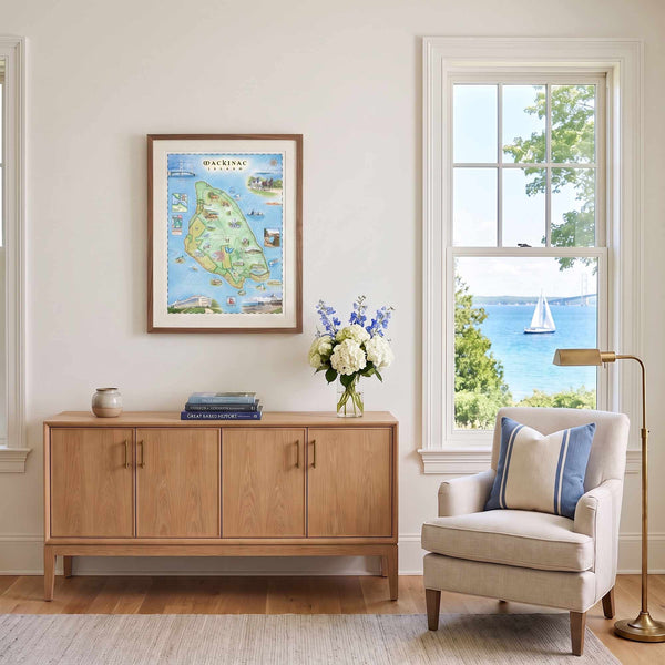 A framed art print of a colorful map of Martha's Vine Island, Virginia, hangs on a cream wall in a beautifully styled living room above a light wood credenza. The room features a beige armchair with blue and white striped pillows, a brass floor lamp, and a window looking out onto a scenic blue ocean with a sailboat.