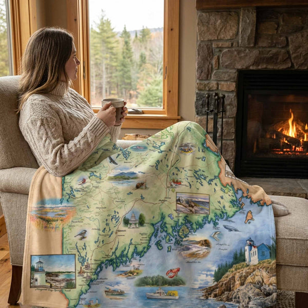 Woman sitting by a fireplace with a scenic blanket draped over her, holding a mug.