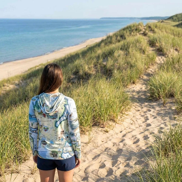 Person standing on a sandy path with grassy dunes and ocean view wearing a sun shirt hoodie