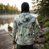 Person wearing a map-patterned hoodie by a lake with trees in the background