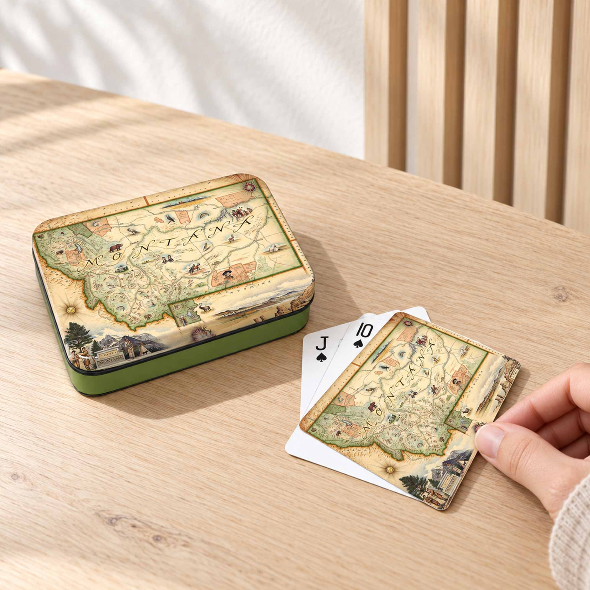 A vintage-style illustrated map tin and matching playing cards on a light wood table, with a hand holding one of the cards. The tin has a green base and the cards show an antique map design with playing card suits visible.