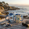 Two mugs with scenic designs on a wooden deck overlooking a coastal landscape.