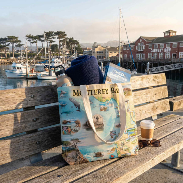 Tote bag with Monterey Bay design on a wooden bench by a waterfront with boats and buildings.